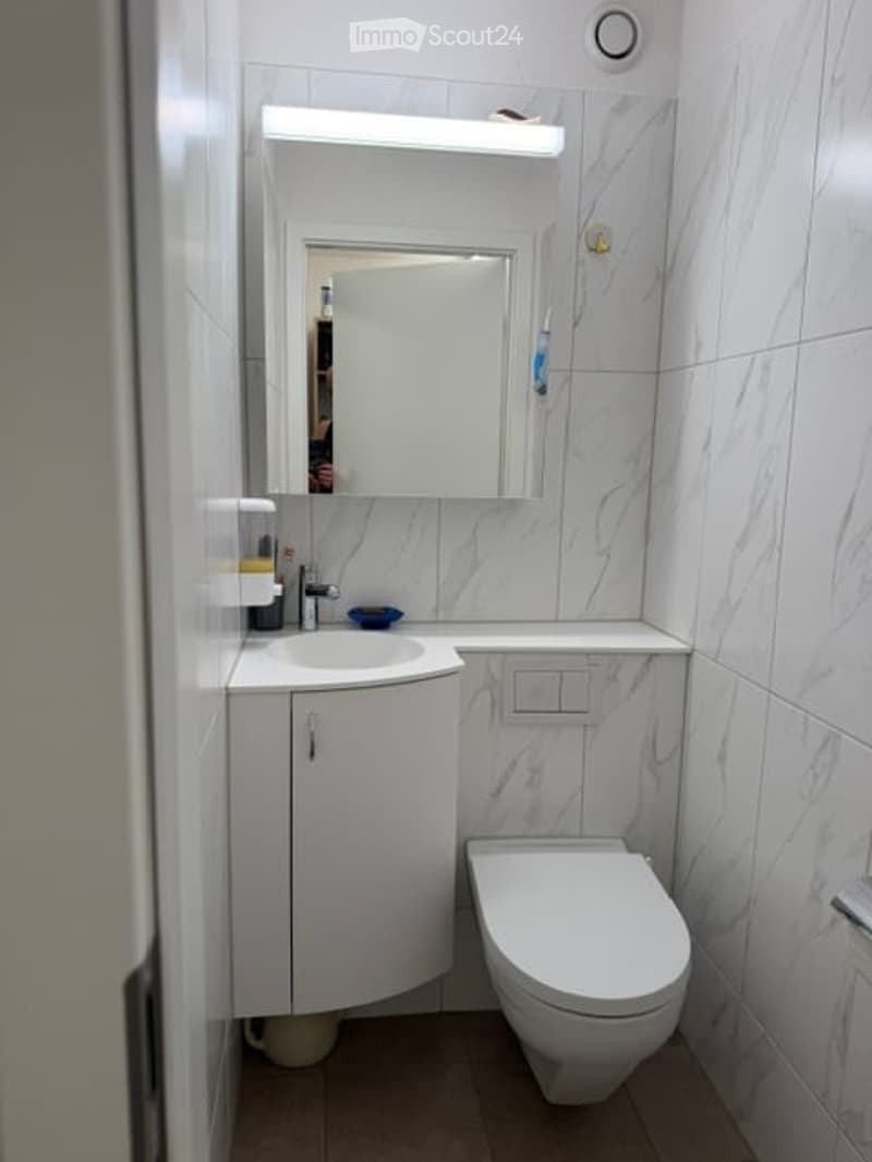 Modern, compact bathroom with white marble tiles, a corner sink cabinet, a toilet, a mirror with built-in lighting, and a toothbrush holder.