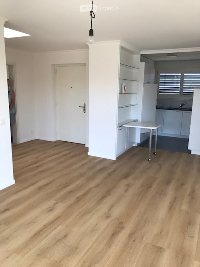 Empty room with light wooden flooring, white walls, a white door, and a small white cabinet with glass shelves. The adjacent kitchen area is visible with white cabinets and a countertop.