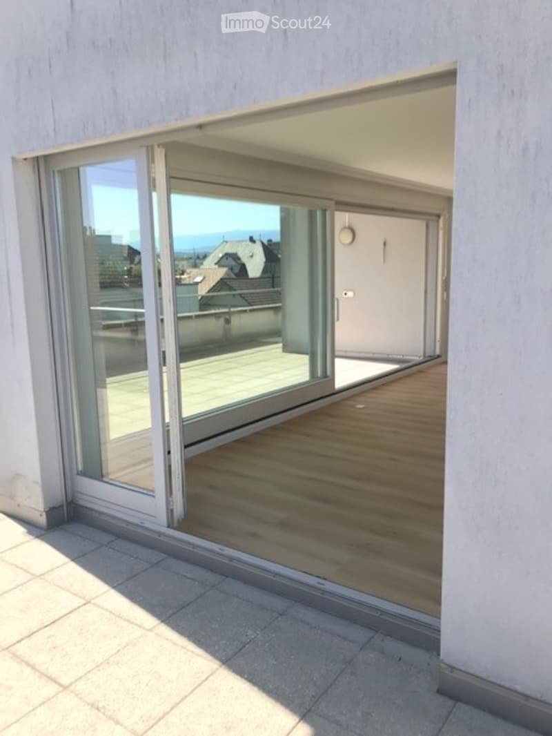 Modern balcony with large glass sliding doors, wooden flooring, and a view of the city and rooftops.