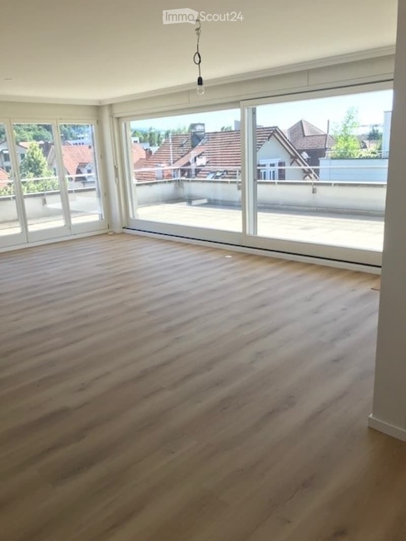 Bright, empty room with large floor-to-ceiling windows offering a view of rooftops and greenery. Light wooden flooring and white walls.