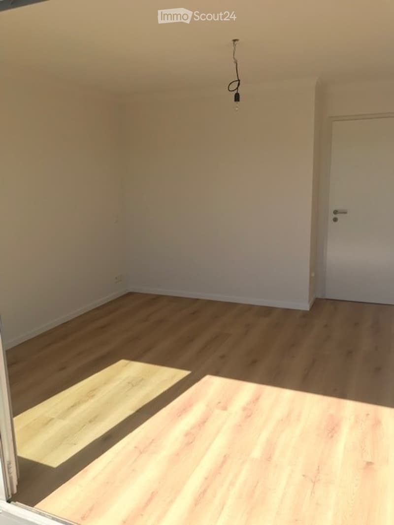 Empty room with light wooden flooring and white walls. A single door is visible on the right side, and a small section of a different flooring type is visible in the foreground.