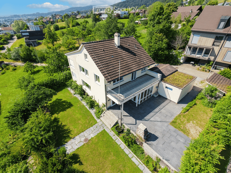 Aerial view of a two-story house with a brown tiled roof, white walls, and a spacious balcony. The property is surrounded by a well-maintained lawn and trees, with neighboring houses visible in the background.