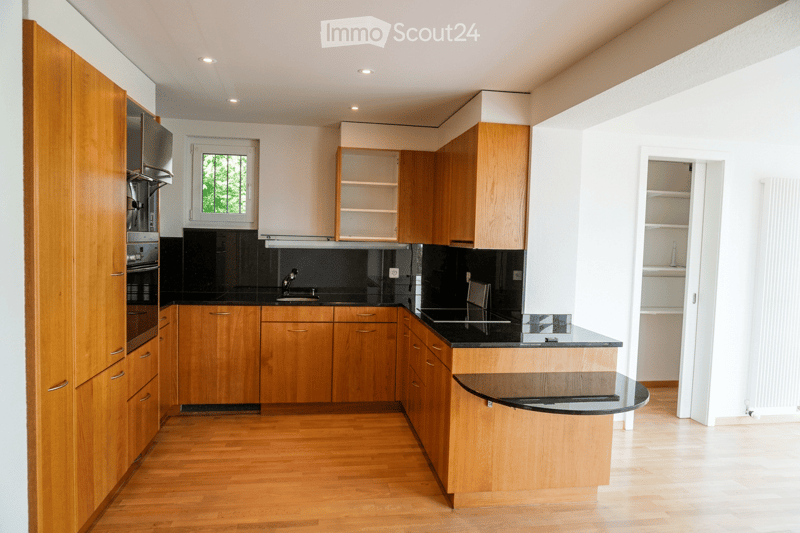 Modern kitchen with light wooden cabinets, black countertops, and stainless steel appliances. Features an island with a sink and a window overlooking greenery.