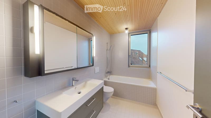Modern bathroom with a white tiled wall, a large mirror with built-in storage, a sink with a modern faucet, a bathtub, and a toilet. The room features a skylight window and a sleek wooden ceiling.