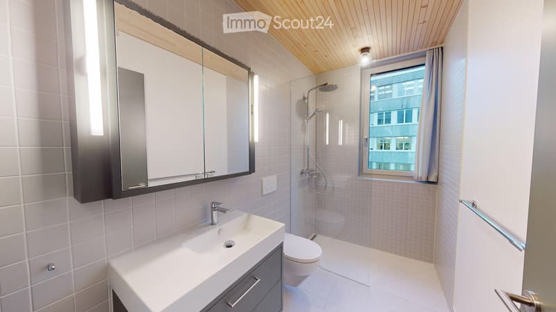 Modern bathroom with white tiles, a large mirror, a sink, a toilet, and a shower. The room features a window with a view of a building outside.