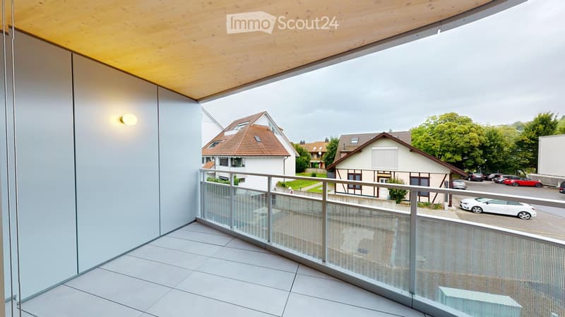 Modern balcony with white tiled floor, glass railing, and wooden ceiling. Offers a view of neighboring houses and parked cars.
