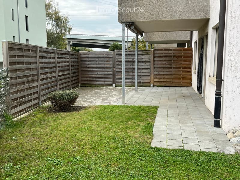 A small, enclosed garden area with a paved pathway leading to a wooden gate, surrounded by a wooden fence and a grassy lawn.