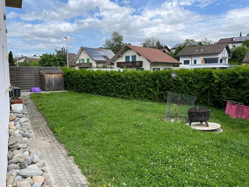 Well-maintained garden with a grassy lawn, a stone pathway, a fire pit, and surrounding hedges. The background shows neighboring houses with solar panels on the roofs.