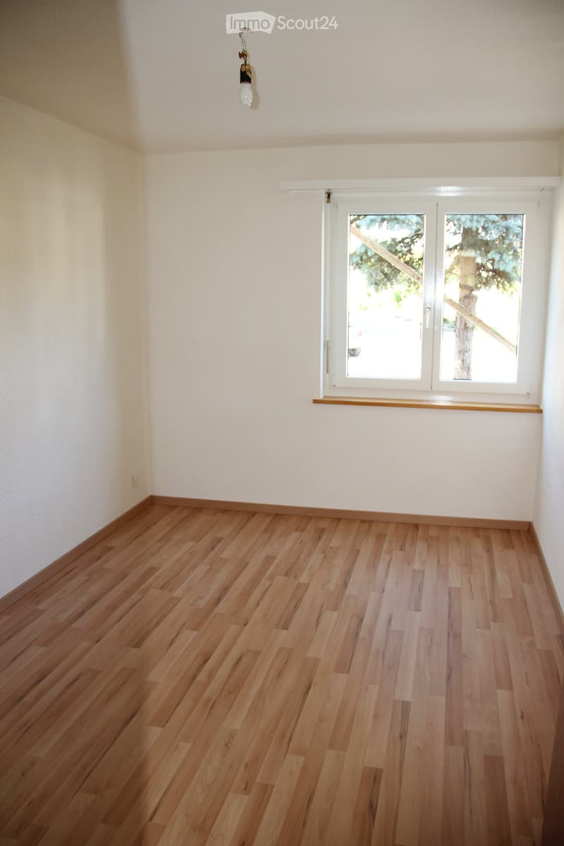 Empty room with light wooden flooring and white walls, featuring a large window with white frames and a small wooden sill.