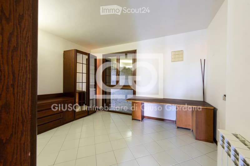 A well-lit room with white tiled flooring, a wooden cabinet with glass doors, a long wooden desk, and a radiator on the wall.