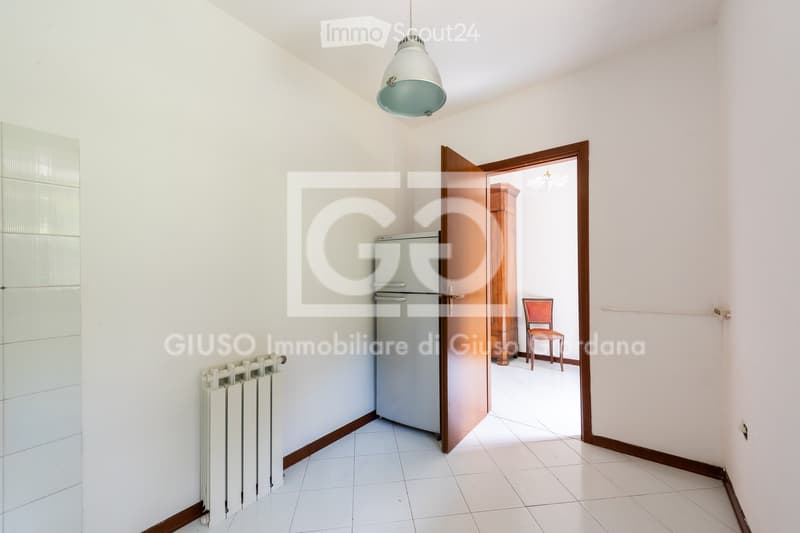 A small, well-lit room with white tiled flooring, a refrigerator, a radiator, and an open doorway leading to another room with a chair visible.
