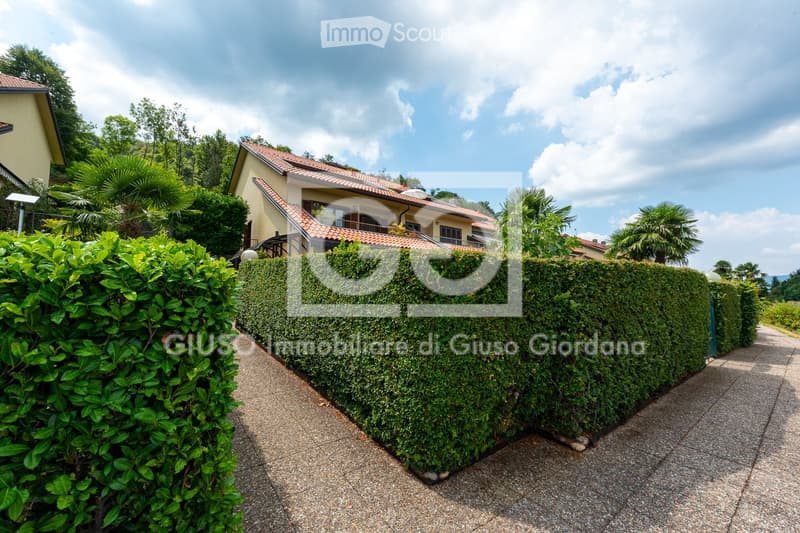 A well-maintained garden path leads to a two-story house with a red-tiled roof, surrounded by lush greenery and trimmed hedges.