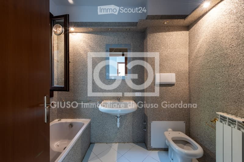 Modern bathroom with a white sink, bathtub, toilet, and a large mirror above the sink. The walls are covered in textured beige tiles, and the floor is tiled in white.