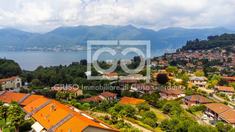 Aerial view of a residential area with numerous houses, orange-tiled roofs, and solar panels, surrounded by greenery and overlooking a lake and distant mountains.