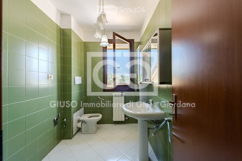 Bathroom with green tiled walls, white tiled floor, toilet, sink, radiator, mirror, and cabinet.