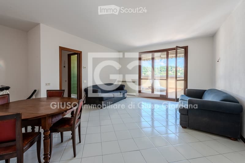 Bright living and dining area with tiled flooring, large sliding glass doors leading to a balcony, a sofa, and a wooden dining table with chairs.