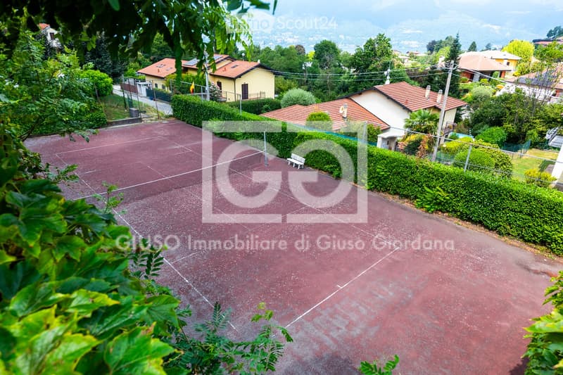 A well-maintained outdoor tennis court with a red clay surface, surrounded by neatly trimmed hedges and trees. The background shows a residential area with houses and a scenic mountain view.