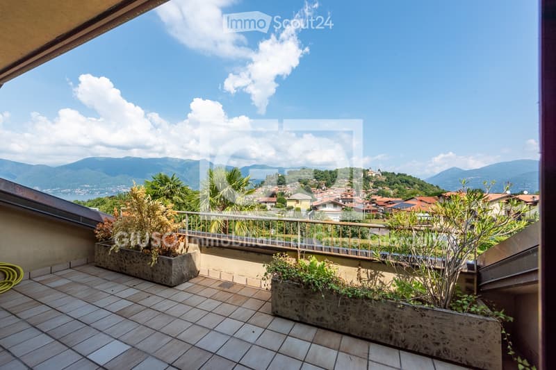 Balcony with tiled flooring, metal railing, and potted plants, overlooking a scenic town and mountainous landscape.
