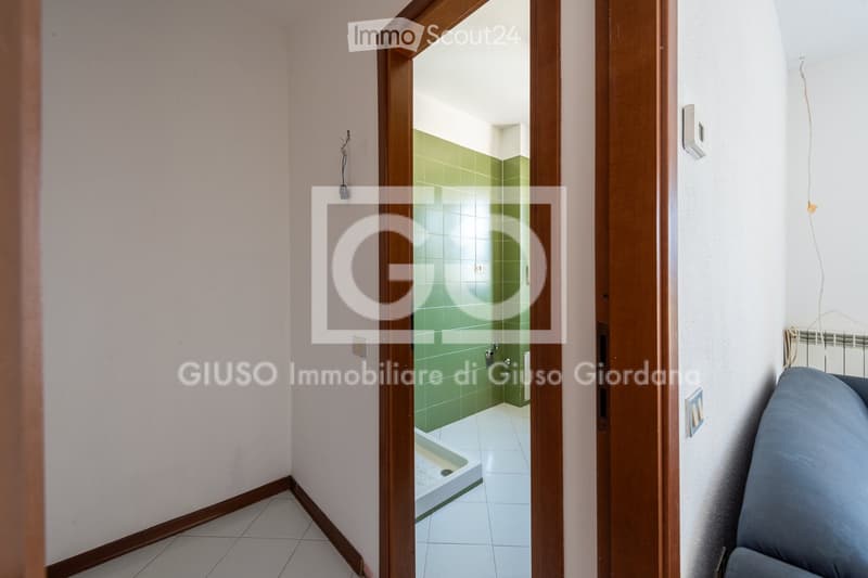 A narrow hallway with light-colored tiled flooring and beige walls, leading to a small bathroom with green tiled walls and a white tiled floor.