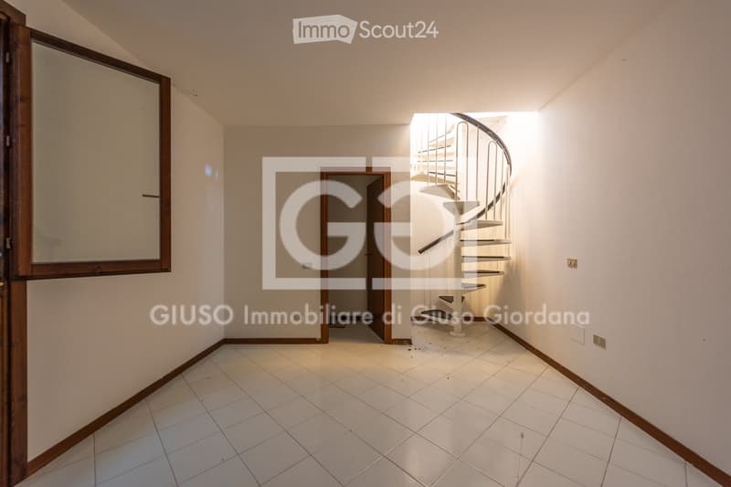 Empty hallway with tiled floor, white walls, and a spiral staircase leading upwards. Features a large mirror on one wall and a door at the end.