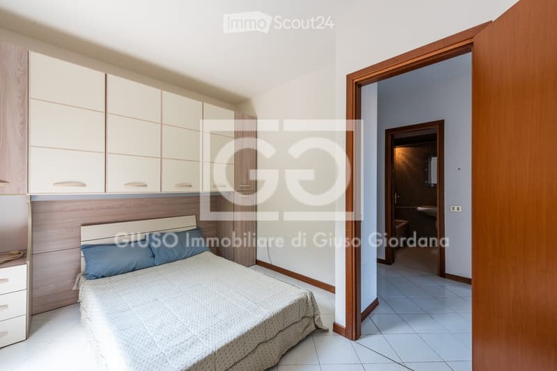 Modern bedroom with a neatly made bed, nightstand, and built-in cabinets. A doorway leads to a bathroom with a sink and mirror.