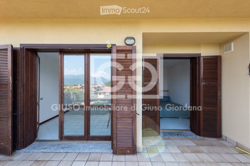 Balcony with large glass doors leading to a tiled outdoor area, overlooking a scenic view of mountains and houses.