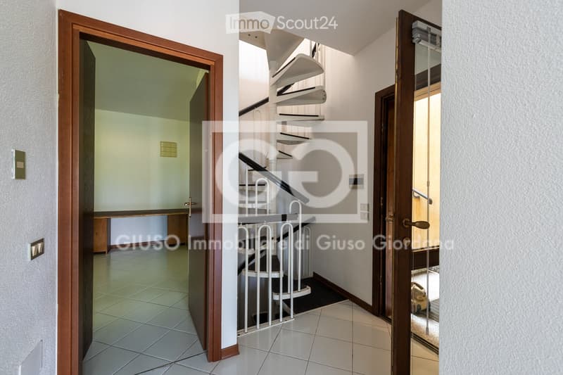 A clean, modern hallway with tiled flooring, white walls, and wooden doors leading to different rooms. A staircase with white railings is visible in the center.