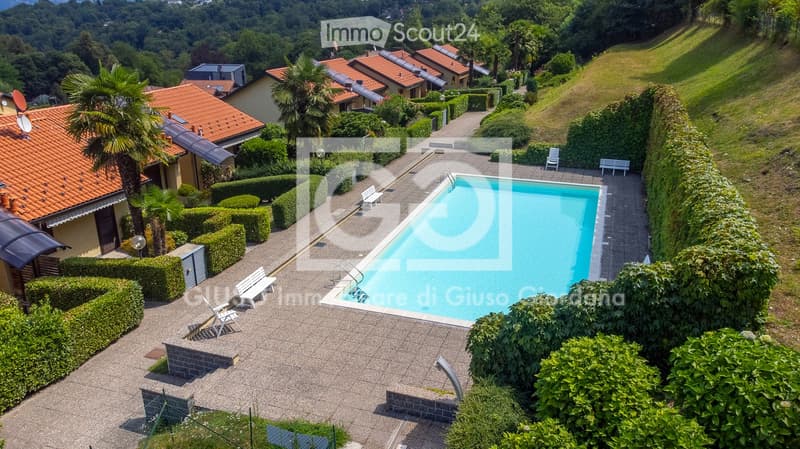 Aerial view of a residential area with a swimming pool surrounded by neatly trimmed hedges, lounge chairs, and benches. The area is bordered by a hedge and includes several houses with red-tiled roofs and satellite dishes.