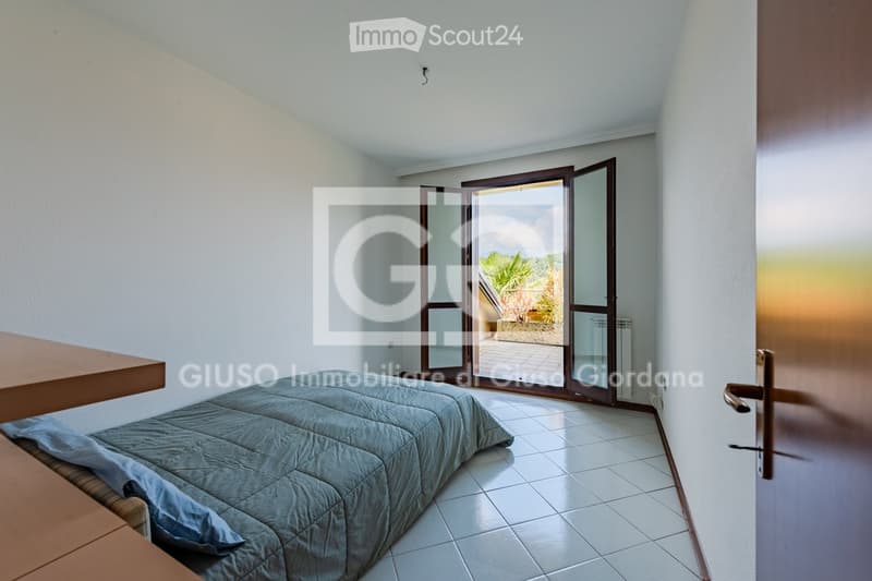 Bright bedroom with a bed covered in blue bedding, a wooden table, and large glass doors leading to a balcony or outdoor area.