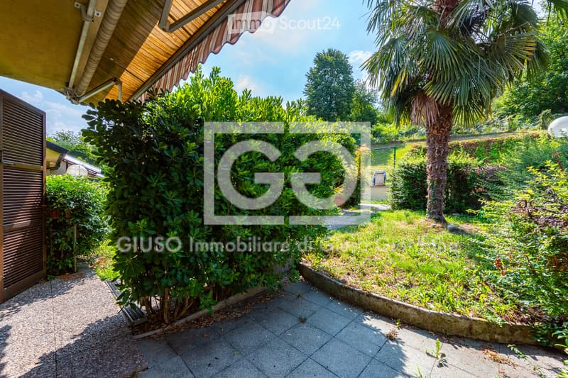 A well-maintained garden area with lush greenery, including bushes, trees, and a palm tree. The space features a paved pathway, a wooden gate, and a covered patio area with a wooden awning.