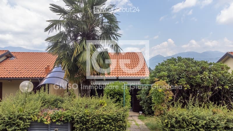 A two-story house with a red-tiled roof, surrounded by lush greenery and a tall palm tree. The house features a covered entrance and is set against a backdrop of mountains.