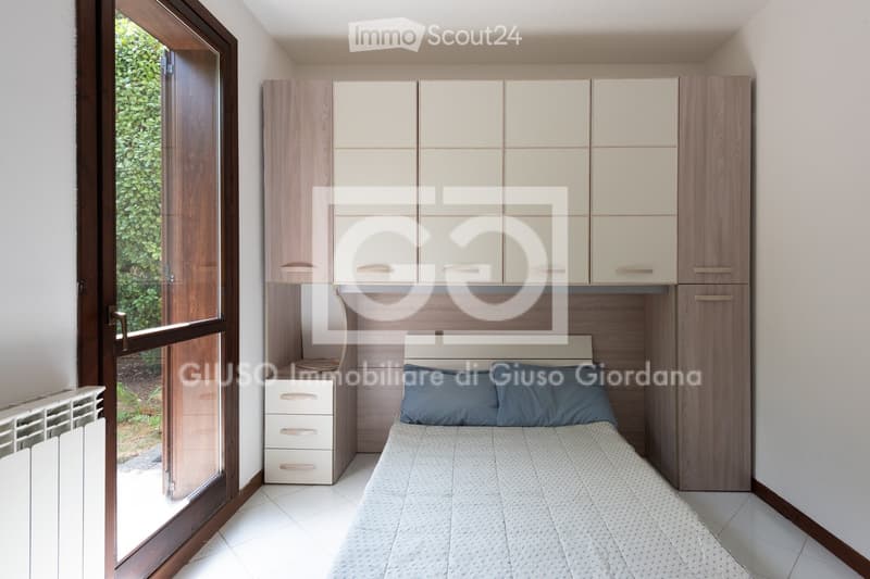 Modern bedroom with a single bed, nightstand, wardrobe, and built-in cabinets. Large window with wooden frame offers natural light.