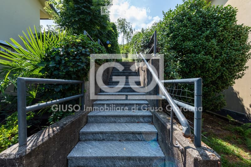 A set of concrete steps with metal railings leading up a raised ground floor, surrounded by greenery and bushes.