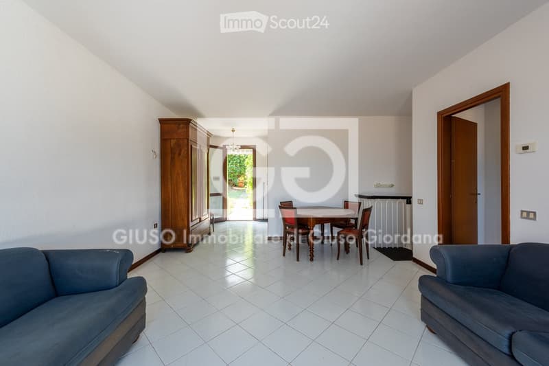 Bright living room with white tiled flooring, two blue sofas, a wooden dining table with chairs, and a large wooden wardrobe. The room has large windows and doors leading to a garden.
