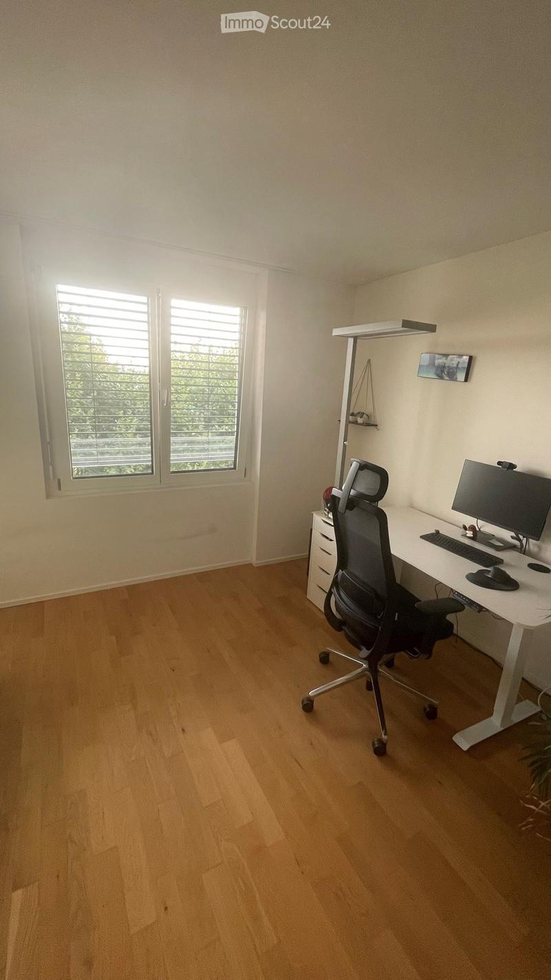 A modern, well-lit office space with a wooden floor, large window with blinds, ergonomic chair, standing desk, computer setup, and small cabinet.