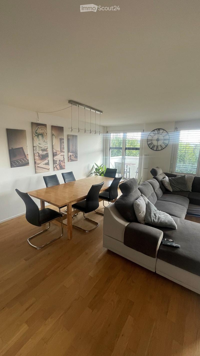 Modern living and dining area with a grey sectional sofa, wooden dining table, chairs, and a large wall clock.