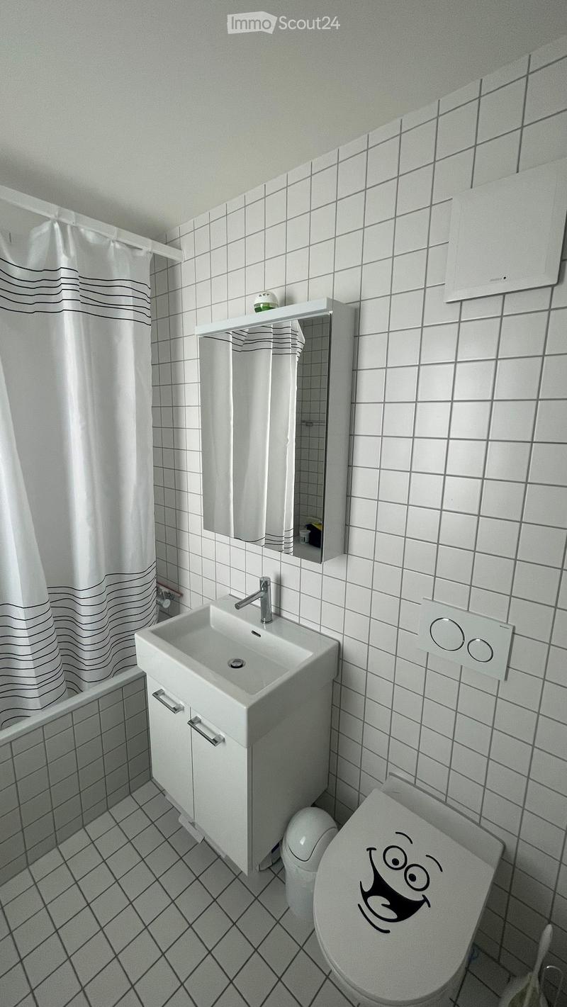 Modern bathroom with white tiles, a sink with a chrome faucet, a toilet with a smiley face sticker, a mirror, and a shower curtain with black wavy lines.