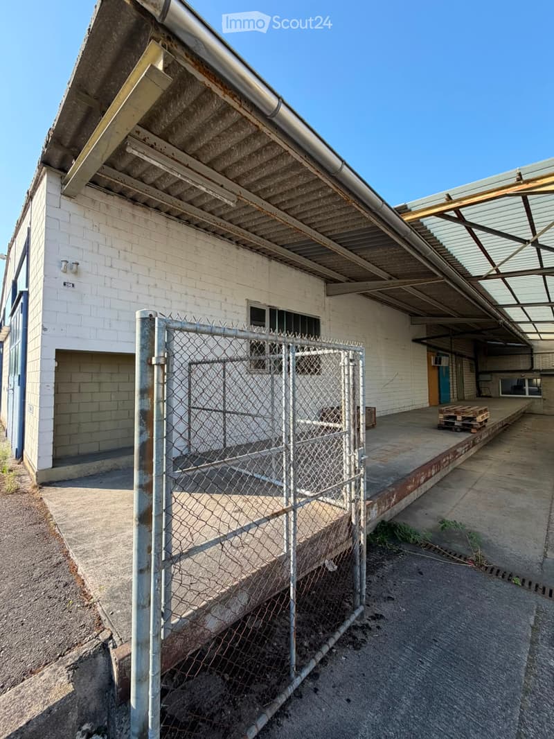 Exterior view of a weathered industrial building with a chain-link fence in front, showing a covered walkway and exposed pipes.