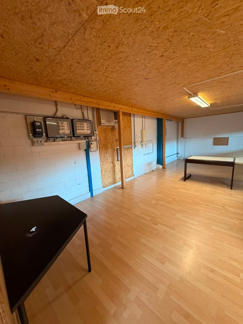 A room with wooden flooring, exposed wooden beams on the ceiling, and a black table in the foreground. The walls are partially exposed brick and partially covered with wooden panels. There is a visible electrical panel and a doorway leading to another roo
