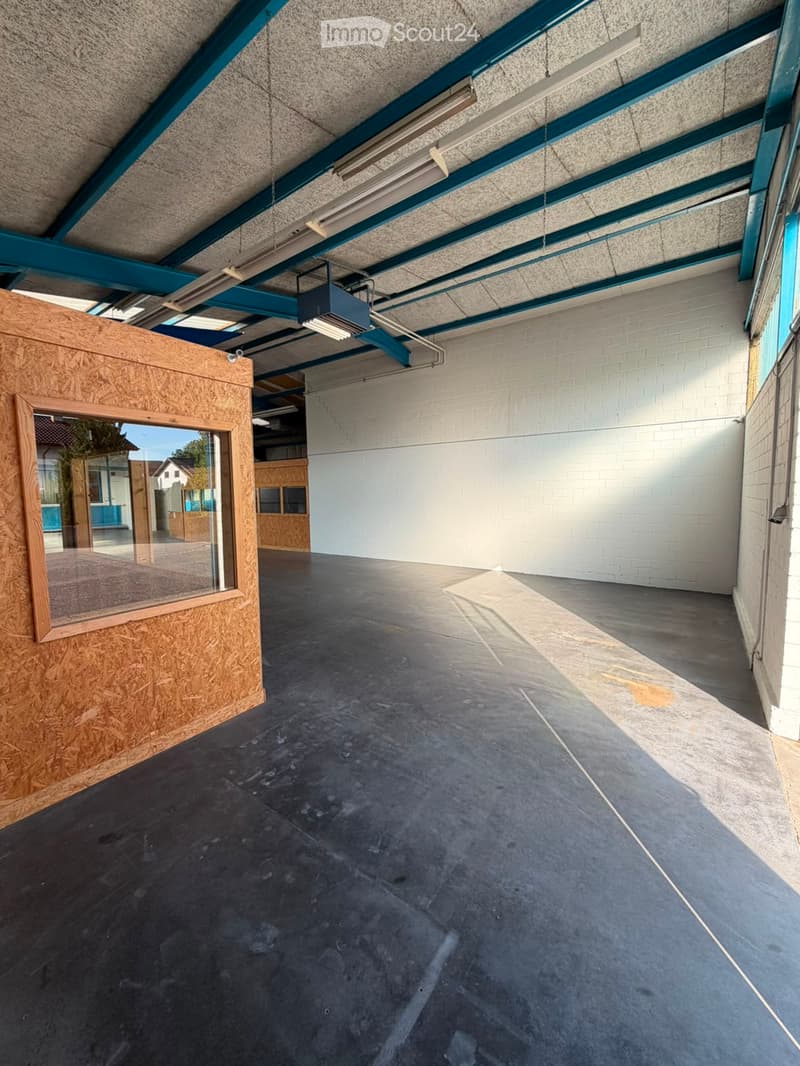 Empty room with concrete floor, white brick walls, and a large window with a wooden frame. The ceiling has exposed beams and fluorescent lighting.