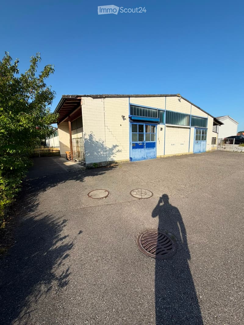 A single-story industrial building with a light-colored facade, blue-framed windows and doors, and a gravel parking area.