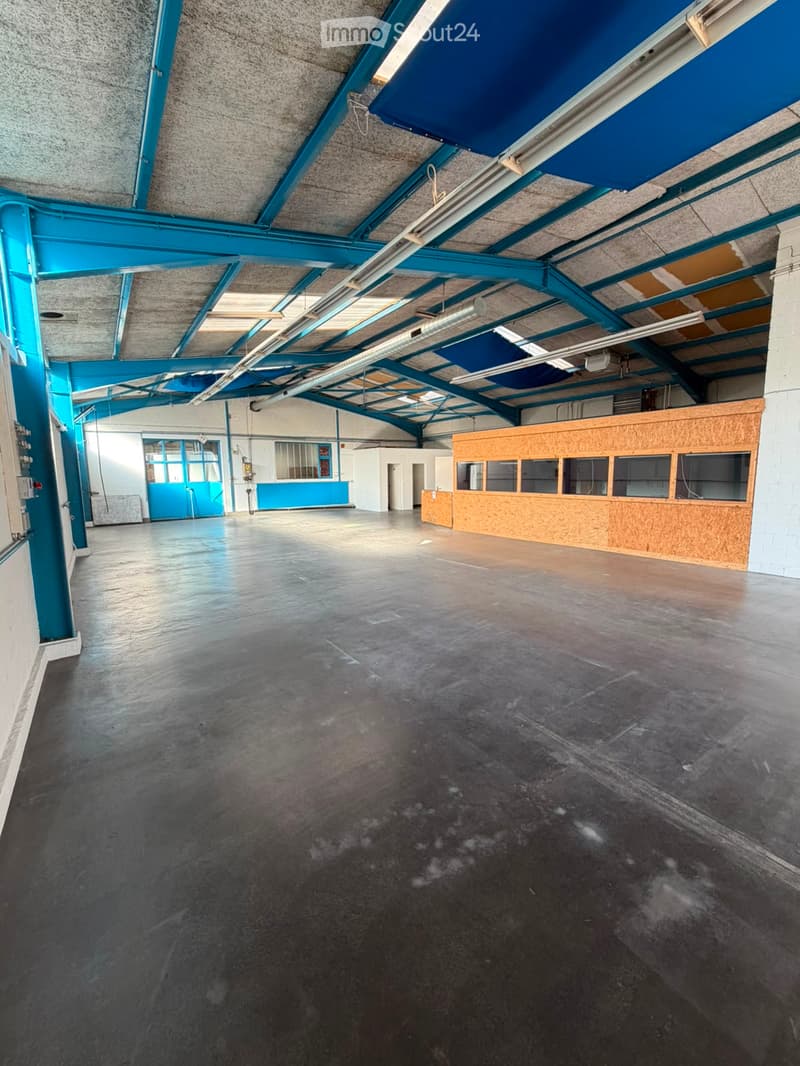 Large, empty industrial-style room with a concrete floor, exposed metal beams, and a wooden reception desk area. The space is well-lit with natural light from windows and has a high ceiling with visible lighting fixtures.