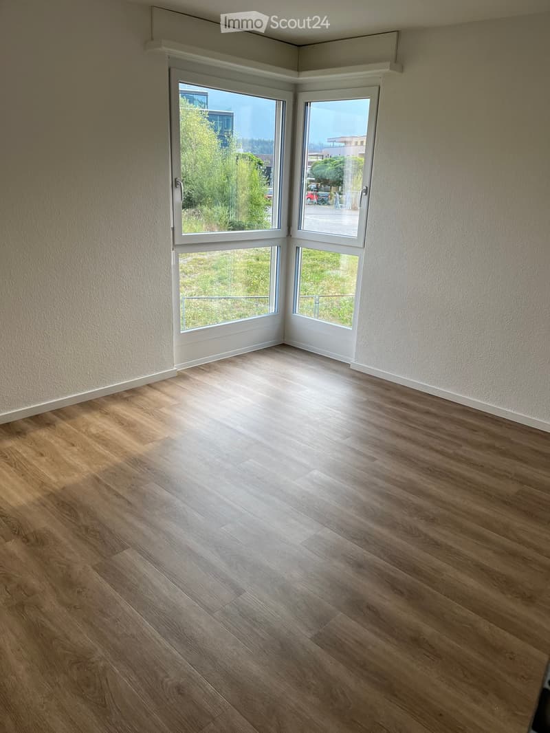 Empty room with wooden flooring and large windows offering a view of greenery and buildings outside.
