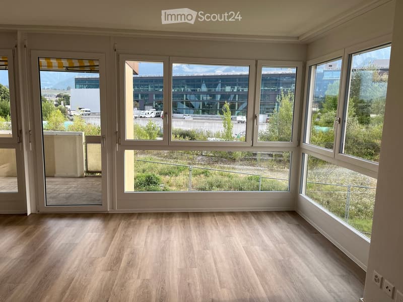 Empty room with light wooden flooring and large windows offering views of the surrounding area, including buildings and greenery.