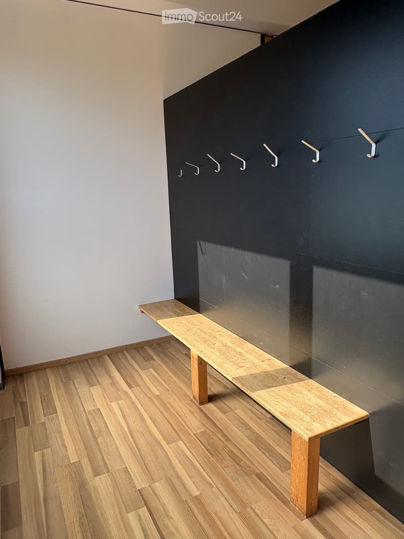 A modern hallway with light wooden flooring, a sleek dark wall with multiple coat hooks, and a simple wooden bench for seating.