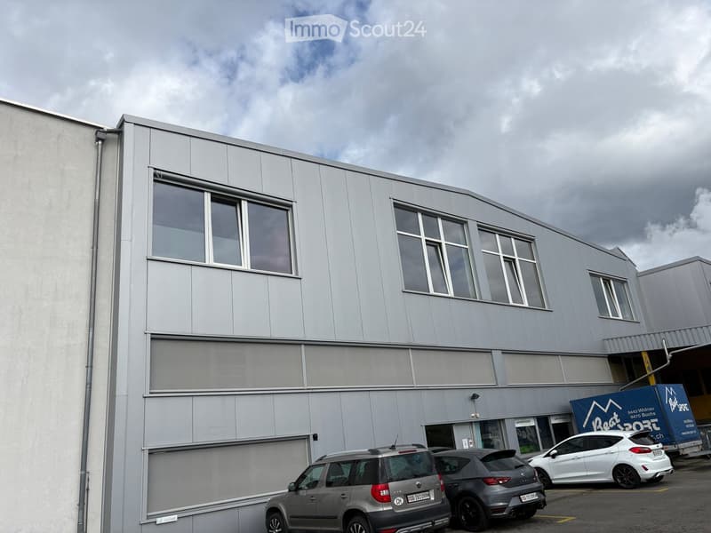 Modern industrial-style building with large windows and a flat roof, featuring several parked cars in front.
