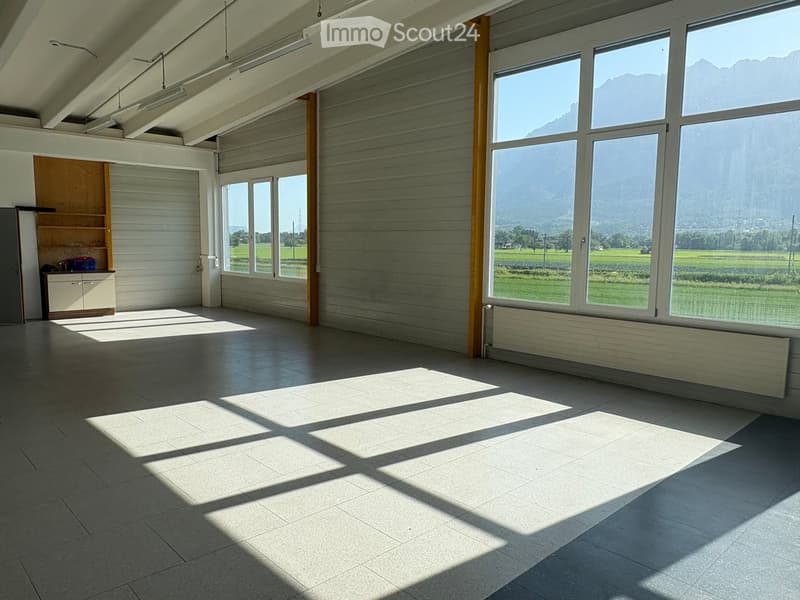 Empty room with large windows, wooden paneling, and a small cabinet with shelves. Natural light streams in, casting shadows on the tiled floor.