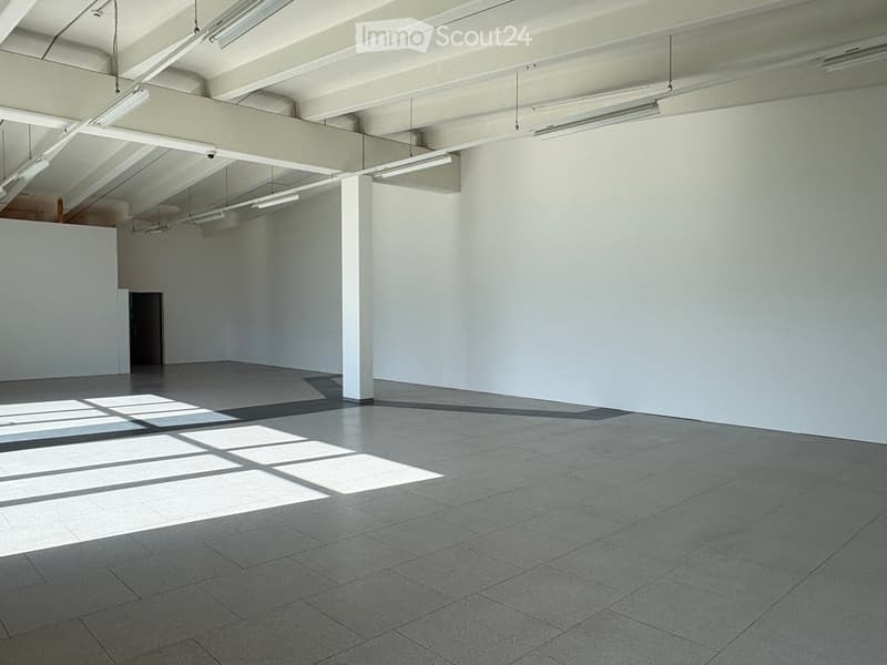 Empty room with white walls, grey tiled floor, and exposed ceiling beams with hanging lights. Sunlight streams in from an unseen window, casting geometric shadows on the floor.