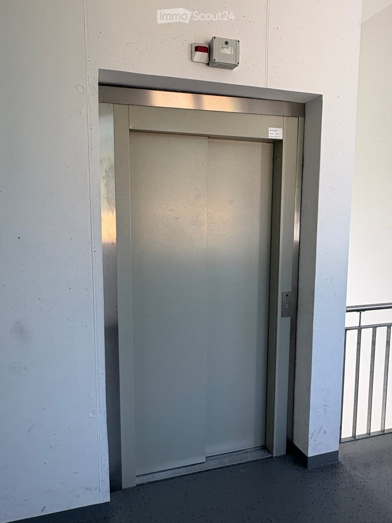 Elevator door in a modern hallway with white walls and a grey floor. A small control panel is mounted above the elevator door.