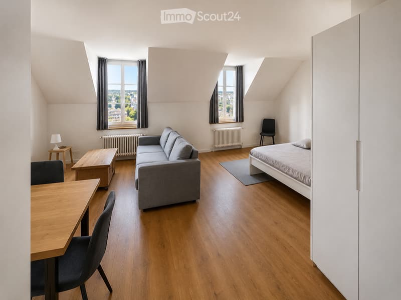 A modern, well-lit attic room with wooden flooring, a grey sofa, a bed, a dining table with chairs, and a small side table with a lamp. The room features large windows with grey curtains and radiators beneath them.
